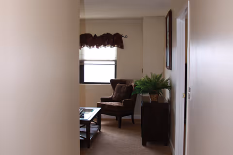 Hallway leading to a small living area with an armchair, coffee table, side table with a potted plant and a window.