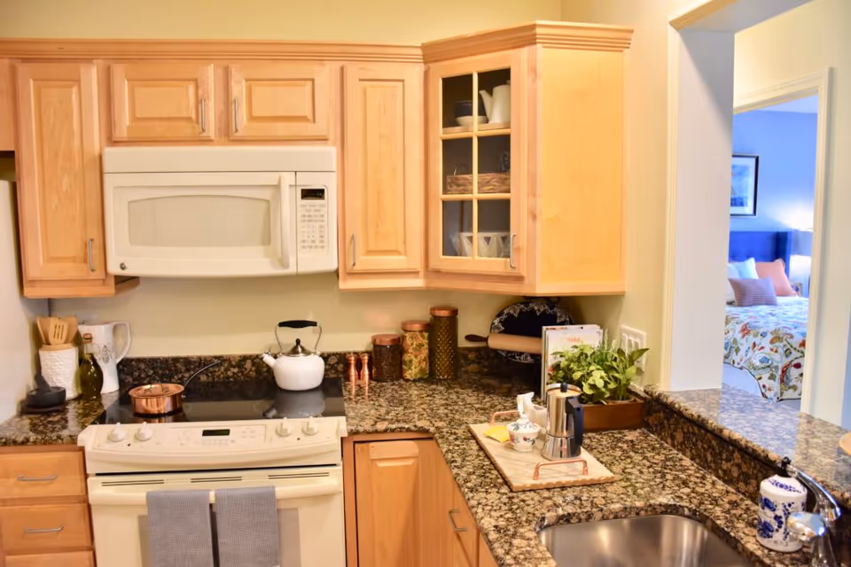 Well-lit kitchen with light wood cabinets, a white microwave and stove, granite countertops and sink, and a pass-through opening to a bedroom.