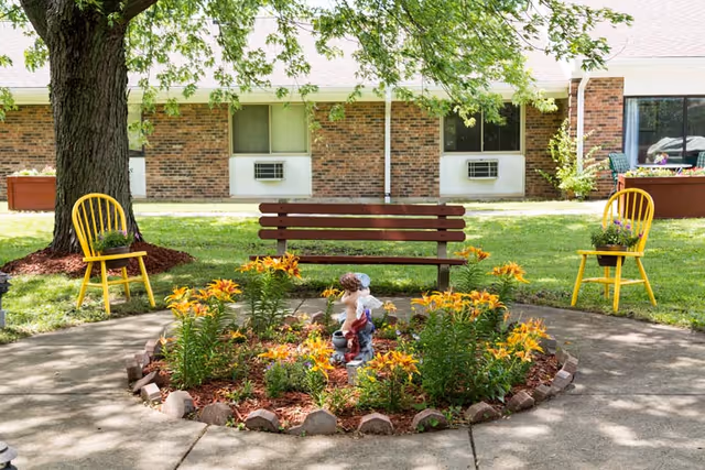 A peaceful outdoor garden area at Cedarview Rehabilitation & Nursing Care featuring a circular flower bed with yellow lilies and a small statue in the center. Surrounding the flower bed is a concrete walkway, a wooden bench, and two yellow chairs with potted plants on them. A large tree provides shade, and a brick building with windows and air conditioning units is visible in the background.