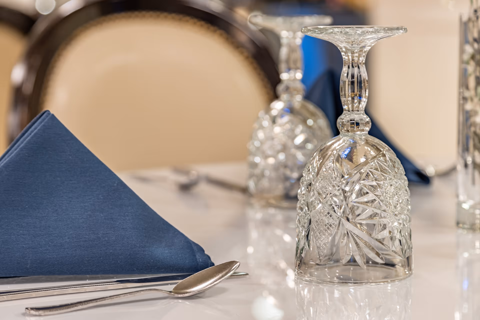 Close-up of a dining table set with an inverted crystal wine glass, a folded navy blue cloth napkin, and silverware including a spoon and knife, with a blurred upholstered chair in the background.