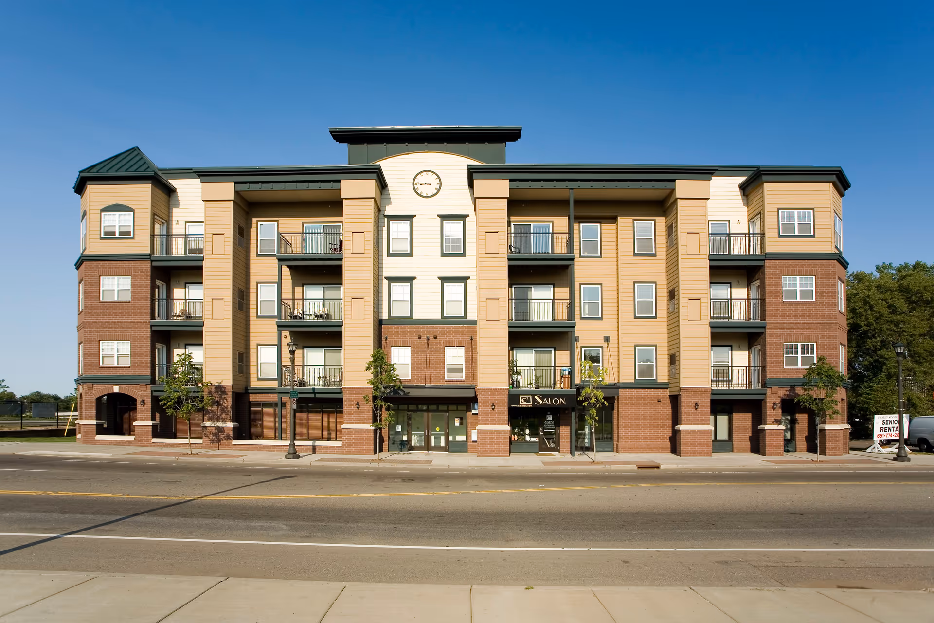 Exterior front view of a four-story senior living apartment building named Phalen Senior Lofts with balconies, a clock above the entrance, and a salon on the ground floor. The building has a mix of brick and beige siding with green trim, and there is a clear blue sky in the background.