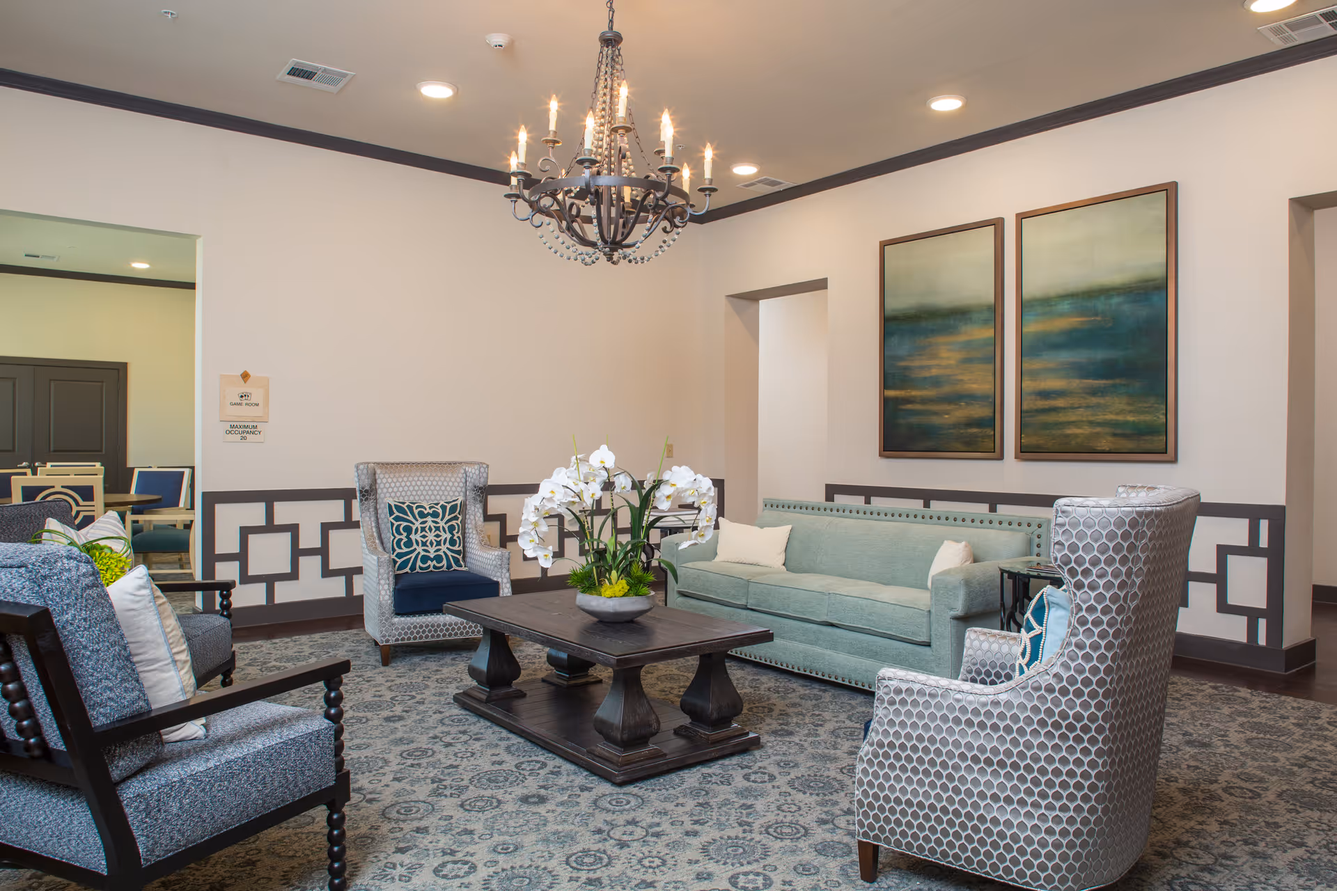 A well-furnished senior living facility common area with a light green sofa, two patterned armchairs, a wooden coffee table with a flower arrangement, and a chandelier hanging from the ceiling. The walls are decorated with two large abstract paintings and have a decorative trim. A dining area is visible through an open doorway.