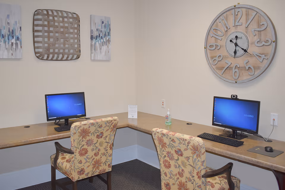 A corner computer workstation area with two desktop computers on a beige countertop. Two floral upholstered chairs are positioned in front of the computers. On the wall above, there is a large round clock and two pieces of abstract wall art with a woven decorative piece in the center. A bottle of hand sanitizer and a small sign are placed on the countertop between the computers.