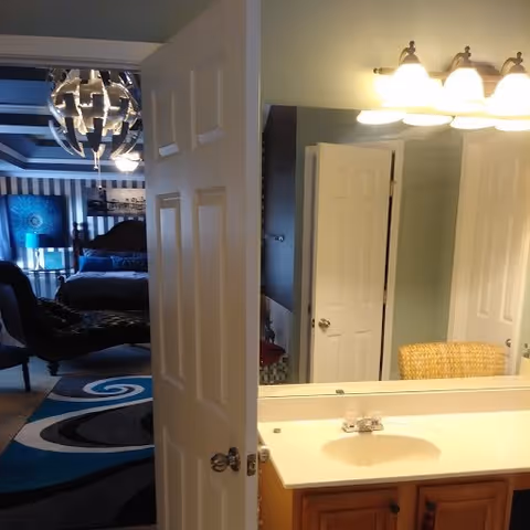View of a bathroom sink and mirror with a partially open door leading to a bedroom. The bedroom features a bed, a chaise lounge, a lamp, and a decorative blue and black rug. The bathroom has a light fixture with multiple bulbs above the mirror.