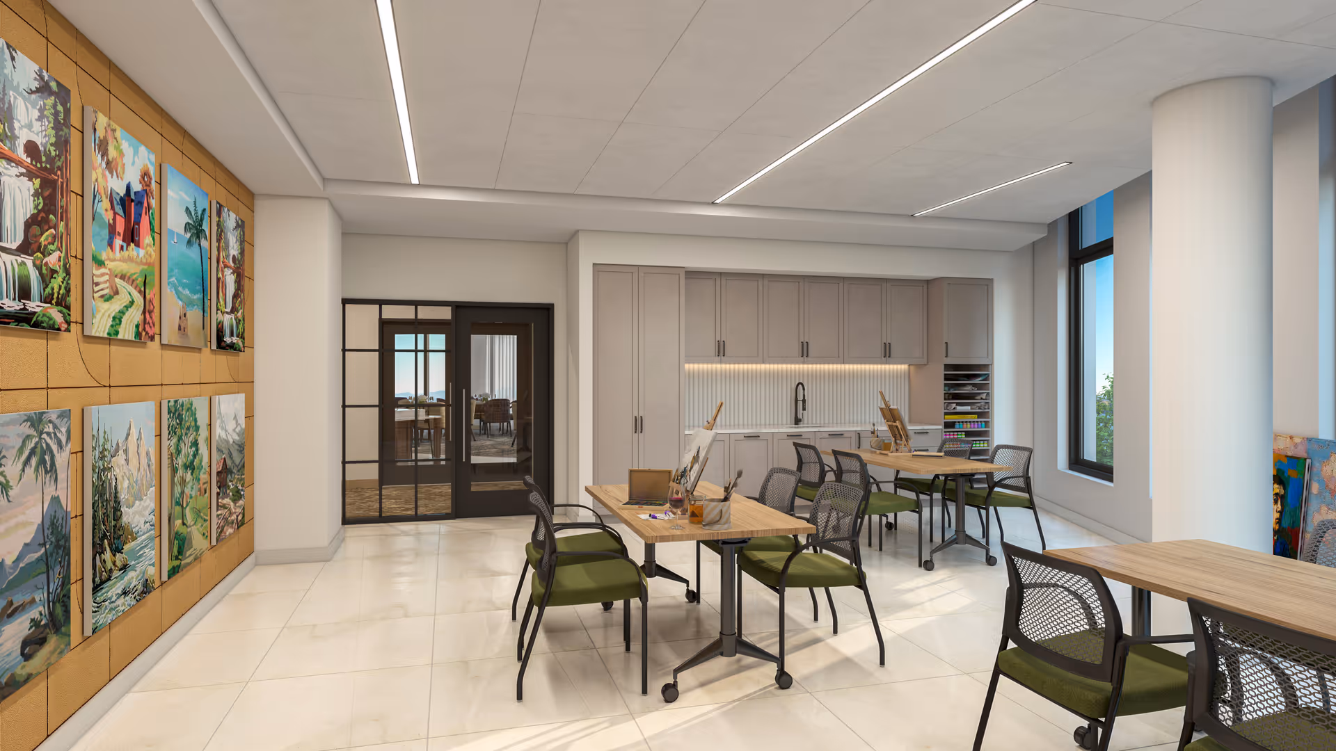 A bright and spacious art room with several tables and chairs arranged for painting activities. The room features a wall with colorful paintings displayed, large windows letting in natural light, and cabinetry with art supplies. The floor is tiled, and there is a double glass door leading to another room.