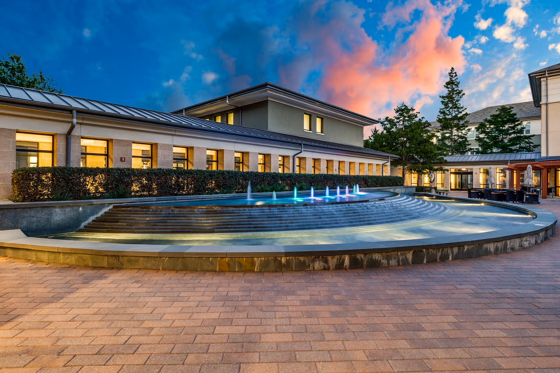A courtyard fountain with colored illuminated jets in front of a senior living building at dusk.