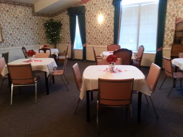 A dining room with several tables covered in white tablecloths, each decorated with a small vase of red flowers. The room has floral wallpaper, dark green curtains, and carpeted flooring. Chairs are arranged around the tables, and there is a large window letting in natural light.
