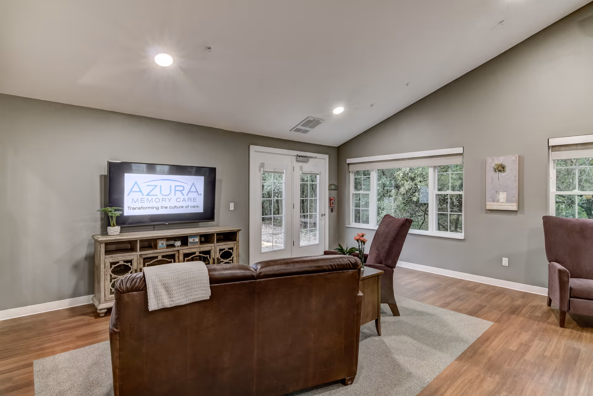Interior common living room with a leather sofa and chairs facing a TV that displays an 'Azura Memory Care' logo, with large windows and wood floors.