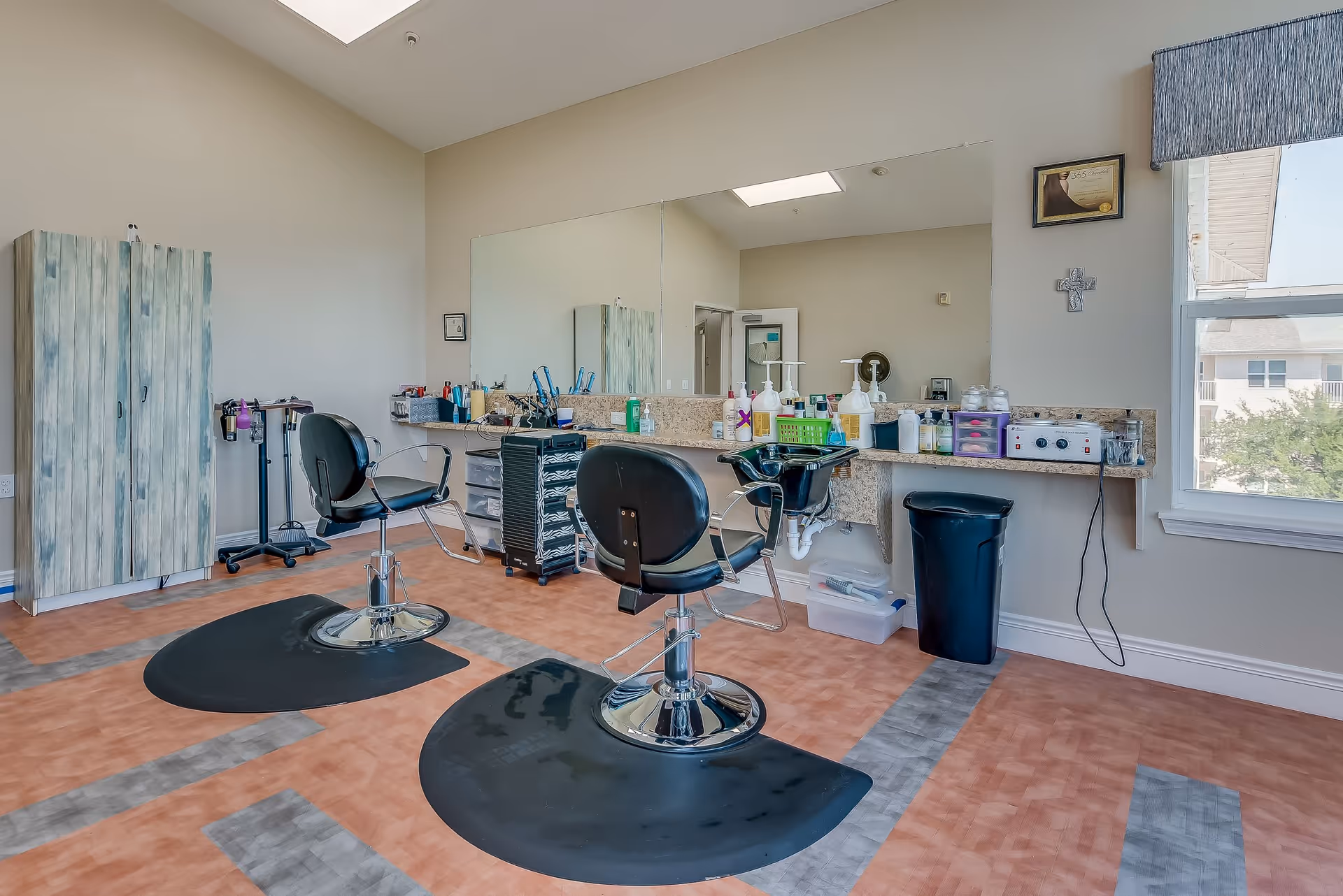 Interior view of a salon area in a senior living facility featuring two black salon chairs on black mats, a large mirror on the wall, a countertop with various hair care products and tools, a cabinet, and a window letting in natural light.