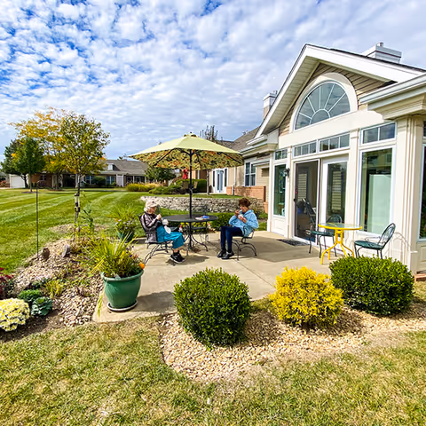 Two elderly people sitting at a round outdoor table under a large green umbrella on a patio next to a building with large windows and a decorative arched window above the door. The patio is surrounded by small bushes and plants, with a well-maintained grassy lawn and other buildings in the background under a partly cloudy sky.