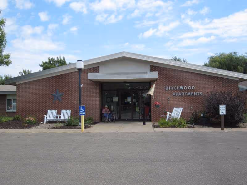 Brick building with Birchwood Apartments sign, entrance doors, benches, and a few residents outside.