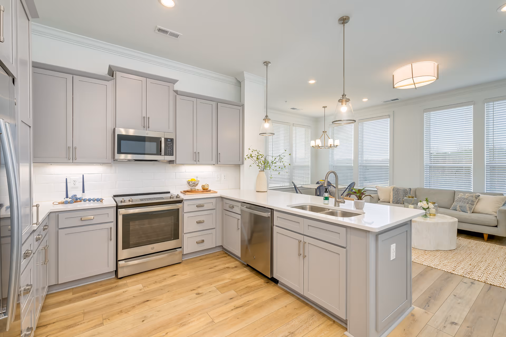 Bright and modern kitchen with light gray cabinets, stainless steel appliances including a microwave, oven, and dishwasher. A large island with a sink and faucet is in the center, decorated with a vase and small plant. The kitchen opens into a living area with a beige sofa, coffee table, and large windows with white blinds letting in natural light.
