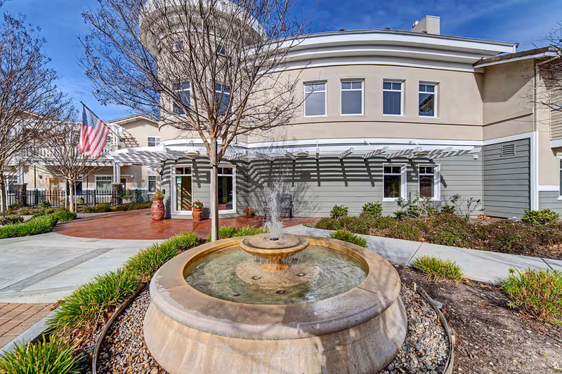 A stone fountain and landscaped entrance in front of the Fremont Hills senior living building with the facade, windows, and an American flag visible.