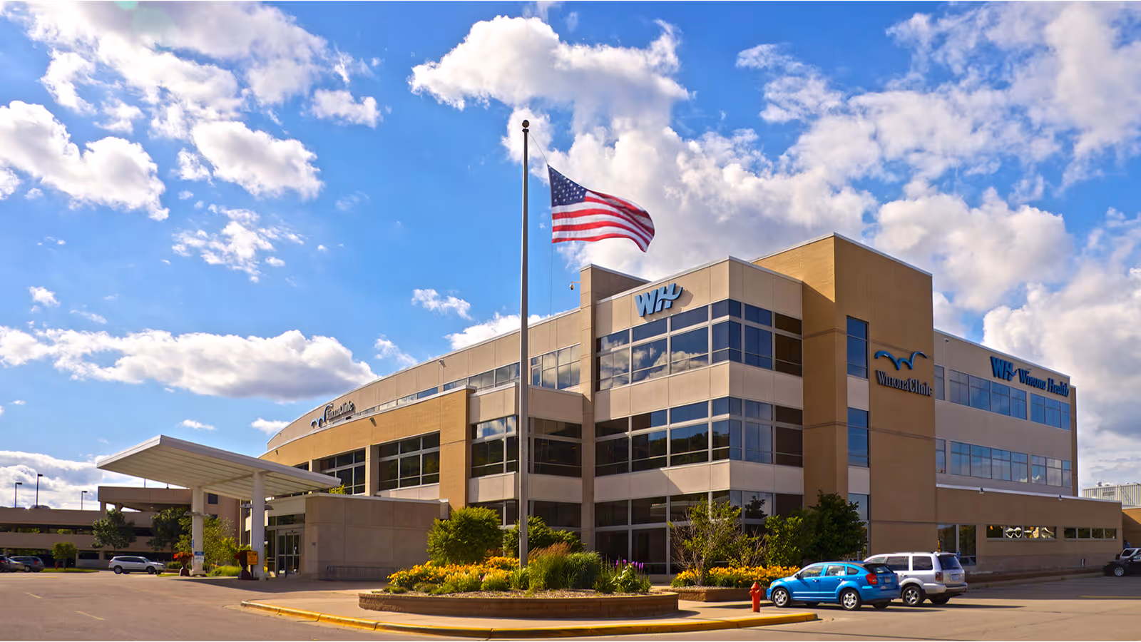 Exterior view of a modern three-story building with large windows under a partly cloudy blue sky. An American flag flies on a flagpole in front of the building, which has landscaped greenery and a circular flower bed near the entrance. Several cars are parked in the parking lot around the building.