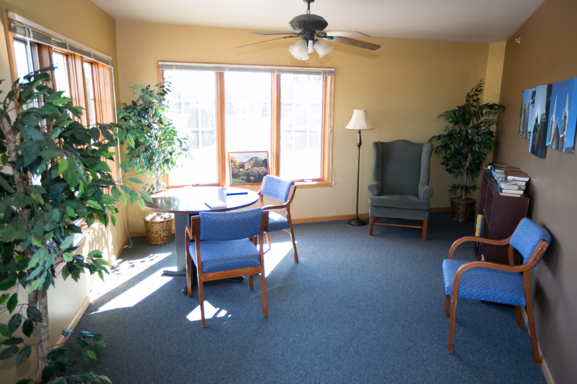 Sunlit sitting room with a small round table, several upholstered chairs, an armchair, potted plants, and a floor lamp.