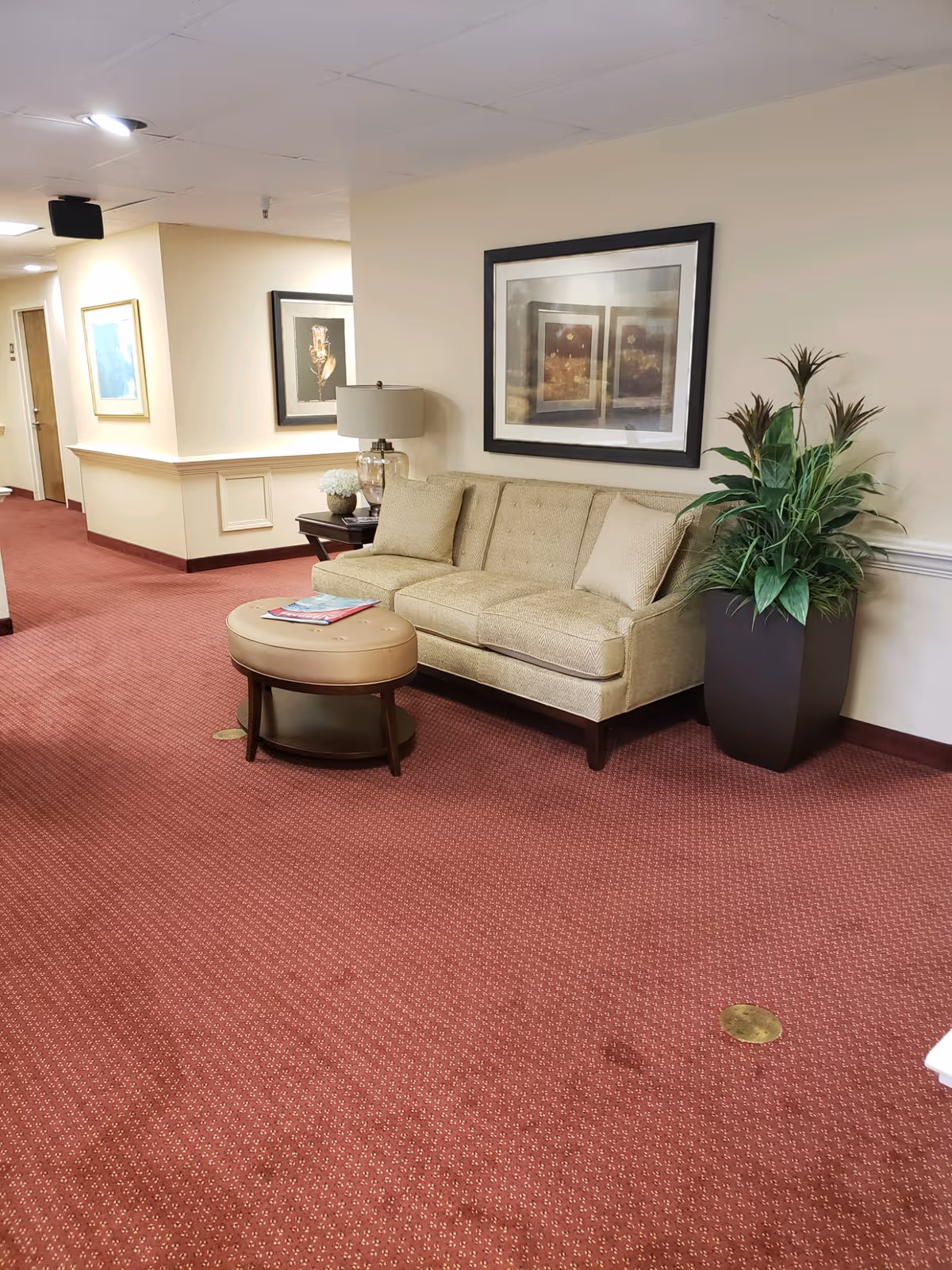 A hallway in a retirement community with red patterned carpet, beige walls, and a beige couch with two cushions. Next to the couch is a round ottoman with magazines on top, a side table with a lamp and decorative items, and a large potted plant. Two framed pictures hang on the walls.