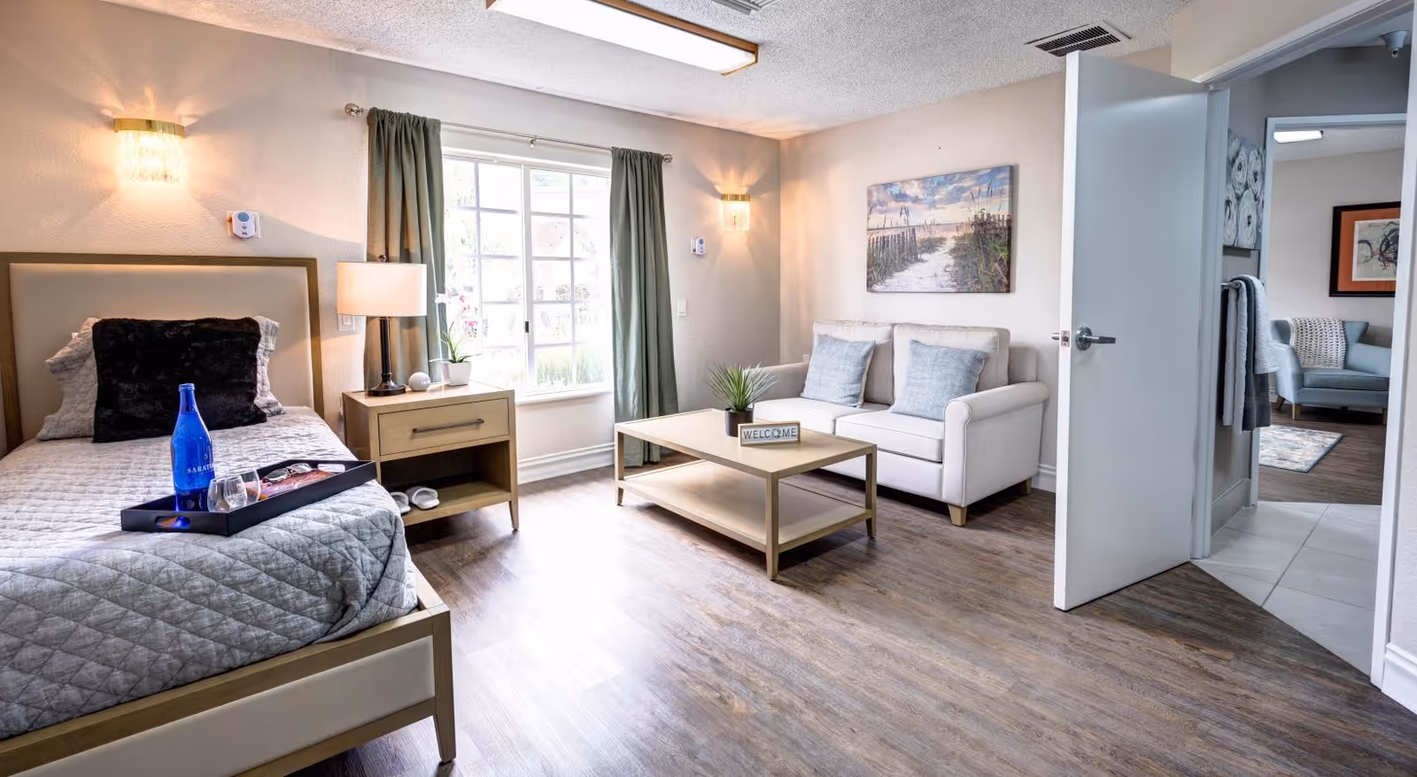 A bright and cozy room in a senior living facility featuring a single bed with a gray quilt and black pillow, a wooden nightstand with a lamp and a small plant, a window with green curtains letting in natural light, a beige loveseat with two gray cushions, a wooden coffee table with a small plant and a welcome sign, and an open door leading to another room with a light blue armchair and framed artwork on the walls.