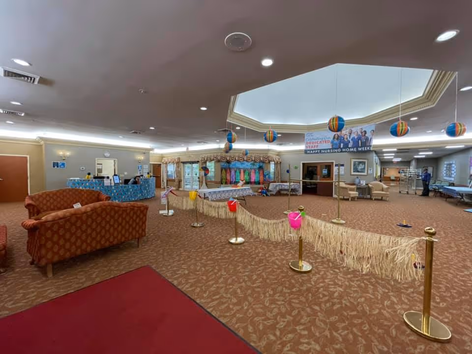 Spacious decorated senior living facility lobby with seating, a reception desk, hanging colorful decorations, and a roped-off activity area.