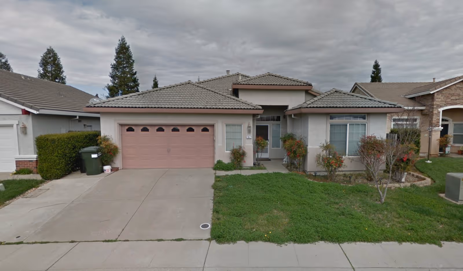 Front exterior view of a single-story house with a tiled roof, a two-car garage with a pink door, a concrete driveway, and a small lawn with shrubs and trees. The sky is cloudy.