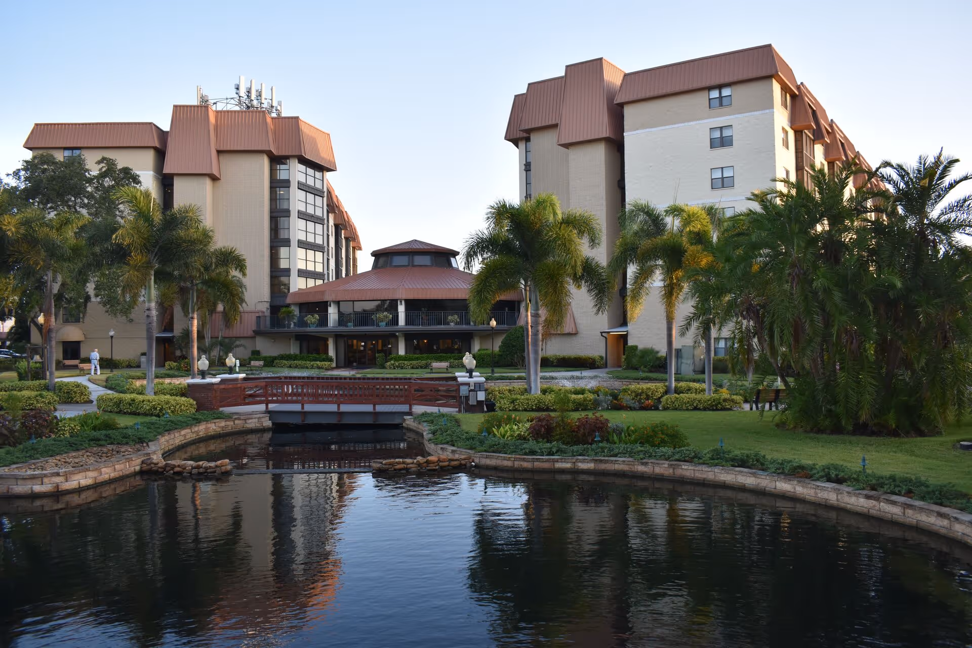 Exterior view of The Inn at Freedom Square, showing a multi-story building with brown roofs surrounded by palm trees and landscaped gardens. A small wooden bridge crosses over a pond in the foreground.