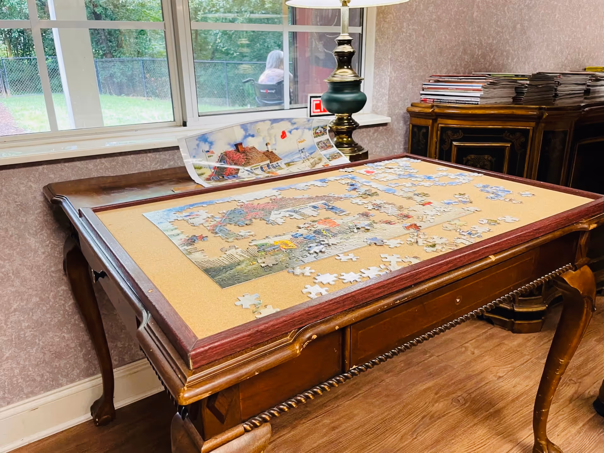 Unfinished jigsaw puzzle spread on a wooden table in a sunlit common room by a window.