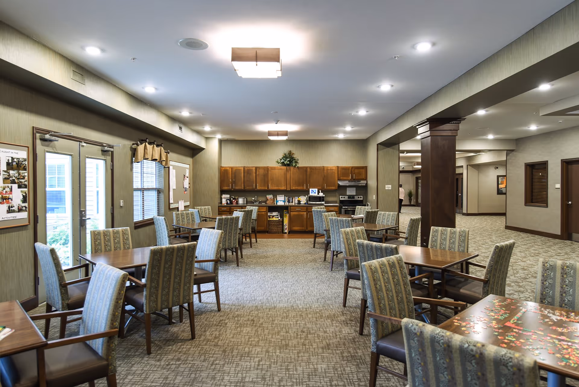 A spacious dining area in a senior living facility with multiple wooden tables and cushioned chairs arranged neatly. The room has a carpeted floor, beige walls, and ceiling lights. At the far end, there is a kitchenette with wooden cabinets, a microwave, and a stove. Large windows and glass doors allow natural light to enter the room.