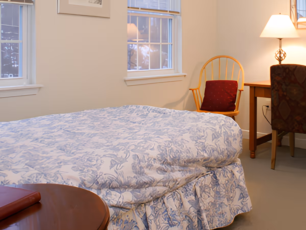 A simple bedroom with a patterned bedspread, a wooden chair with a red cushion, a desk and lamp beside two windows.