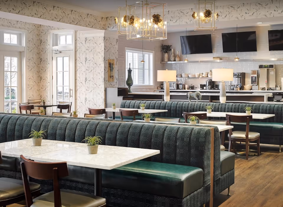A bright and modern dining area with green upholstered booth seating and wooden chairs around white marble tables. Small potted plants decorate each table. The room features large windows and glass doors letting in natural light, floral wallpaper, and pendant lights with decorative bulbs. In the background, there is a counter with kitchen appliances and three mounted flat-screen TVs above it.