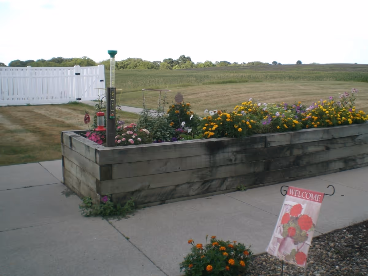 A raised wooden garden bed filled with colorful flowers including yellow, pink, and white blooms. A white fence and a grassy field are visible in the background. A small sign with red flowers and the word 'WELCOME' is placed on the ground near the garden bed.