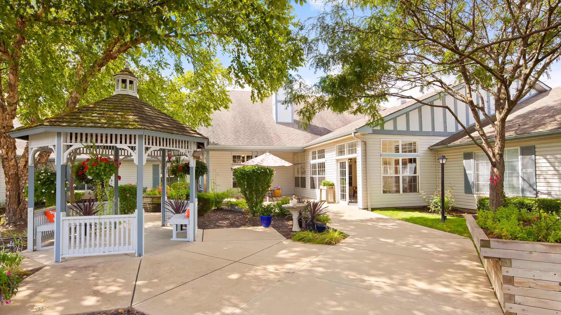 Outdoor courtyard area of a senior living facility with a white gazebo adorned with hanging flower baskets, surrounded by trees and greenery. The building has large windows and a light-colored exterior with a walkway leading to the entrance. There are benches and landscaped garden beds around the courtyard.