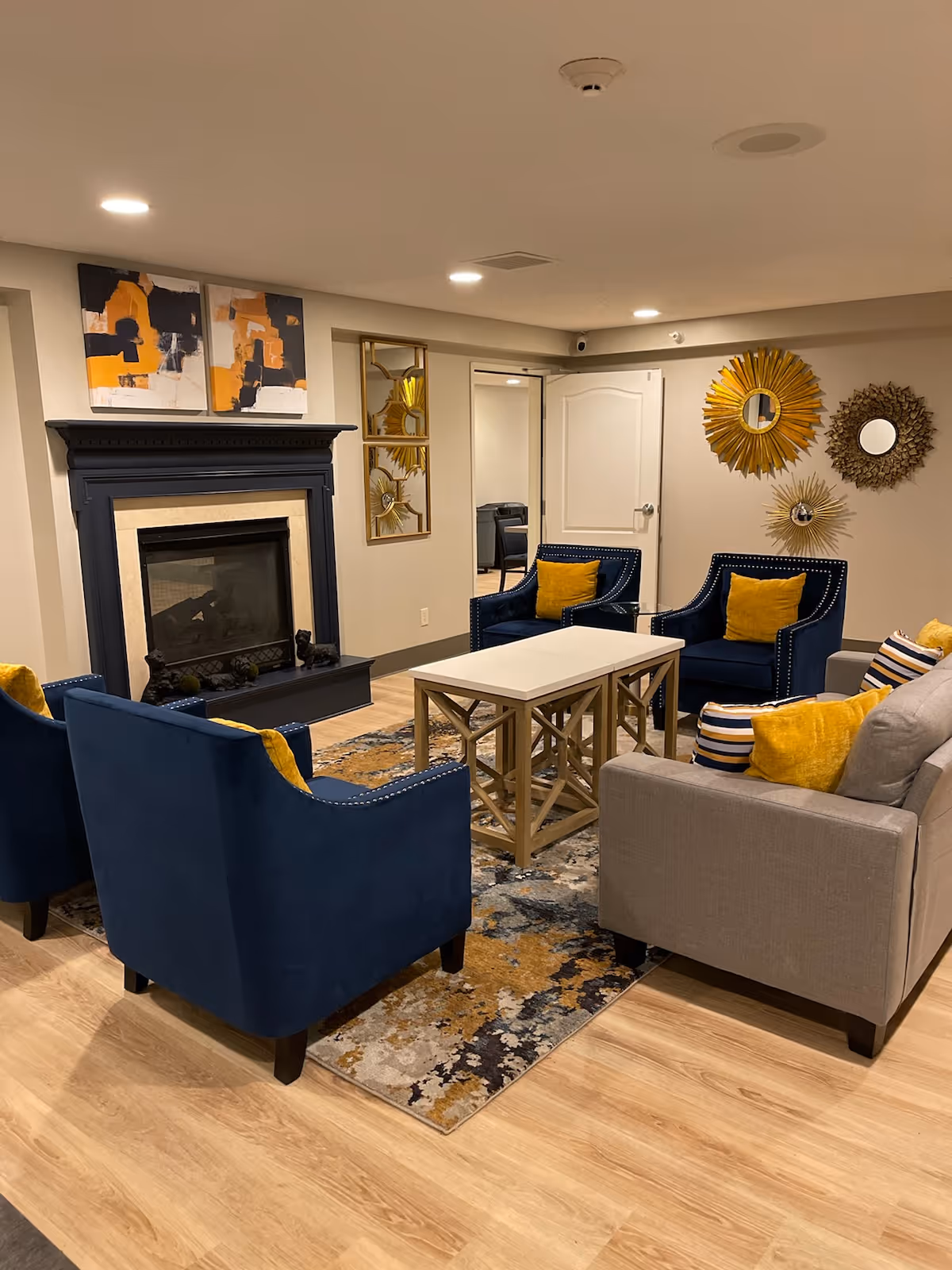 A cozy living room in a senior facility with a fireplace, blue armchairs, a light coffee table, and wall mirrors.