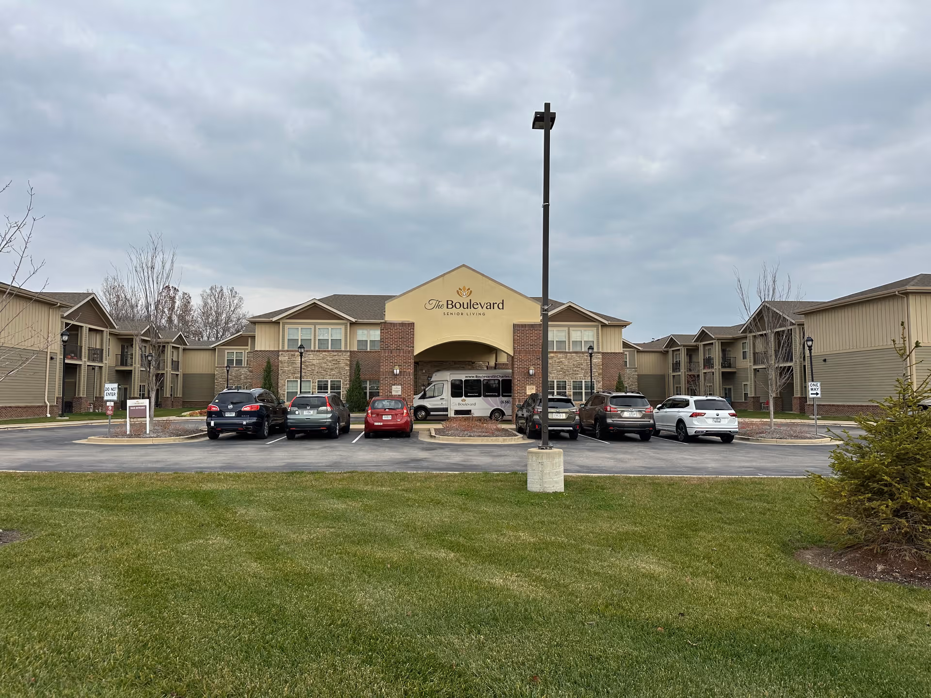 Front exterior view of The Boulevard Senior Living facility in St Charles, showing a two-story building with a parking lot in front, several parked cars, a grassy area, and a cloudy sky.