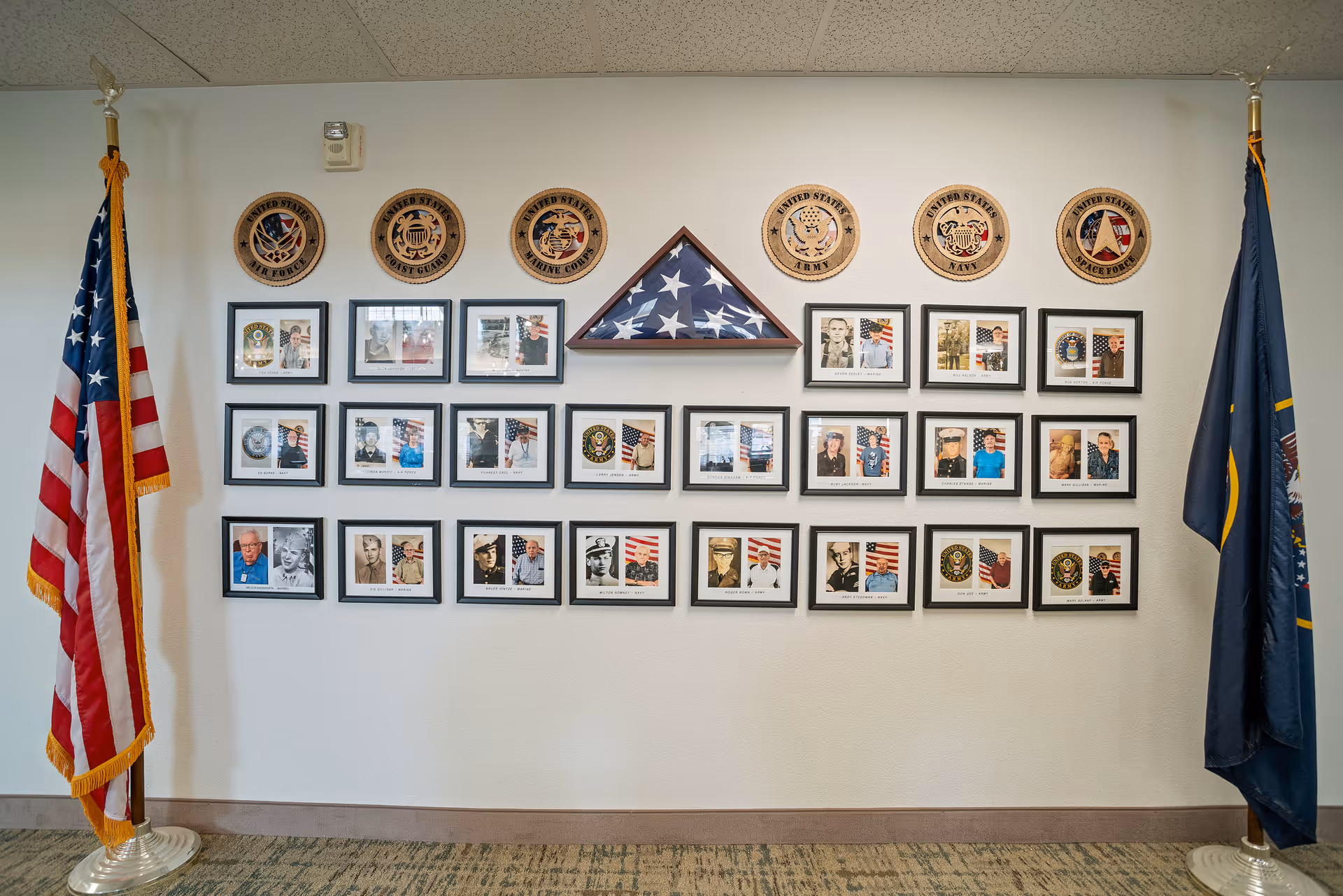 A wall display featuring framed photographs of veterans, military insignias, and a folded American flag in a triangular case. The display is flanked by two flags, one American flag on the left and another flag on the right, in a room with carpeted floor and white walls.
