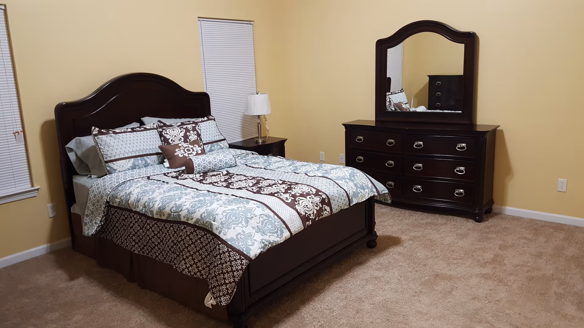 A neatly made bed with patterned bedding, nightstand and lamp, and a dresser with mirror in a carpeted bedroom with yellow walls.