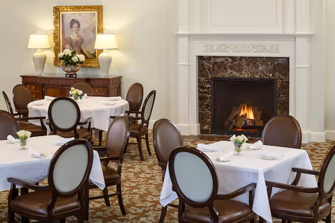 A cozy dining room with round tables covered in white tablecloths, each set with napkins and small flower arrangements. The room features elegant wooden chairs with upholstered backs, a fireplace with a marble surround, and a sideboard with two lamps and a framed portrait above it.