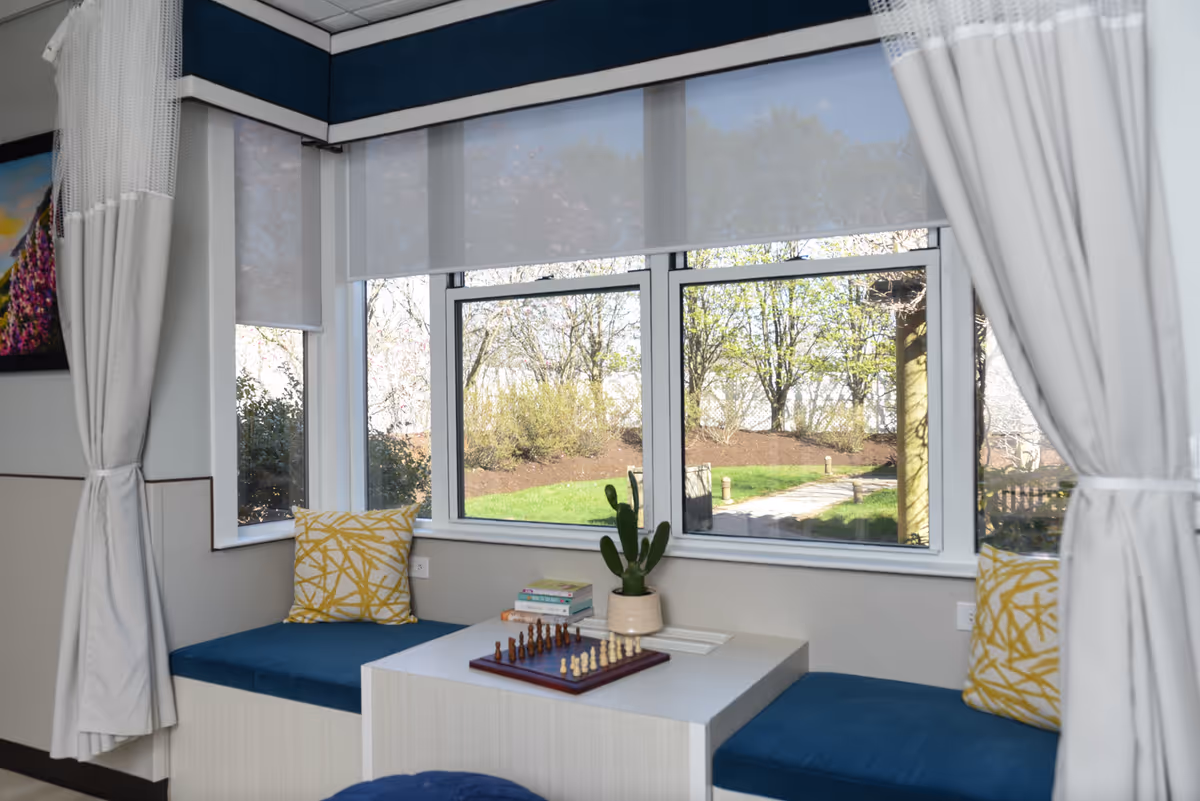 A cozy seating nook with blue cushioned benches and yellow patterned pillows on either side of a white table. On the table, there is a chessboard, a small potted cactus, and a stack of books. Large windows behind the seating area offer a view of a green outdoor space with trees and a pathway. White curtains frame the windows and the area has a calm, inviting atmosphere.