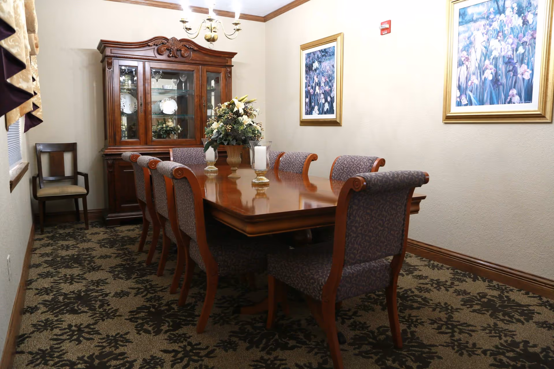 A formal dining room with a long wooden table surrounded by eight upholstered chairs. A floral centerpiece and two candle holders are on the table. Behind the table is a wooden china cabinet displaying plates and glassware. The room has patterned carpet, two framed floral paintings on the wall, and a chandelier hanging from the ceiling.