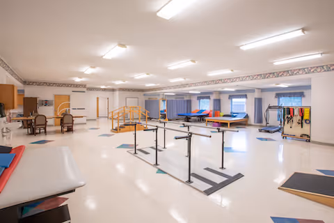 A spacious rehabilitation therapy room with parallel bars for walking exercises, therapy tables, exercise equipment, and chairs. The room has bright overhead lighting, large windows with blue curtains, and a clean, open floor space.