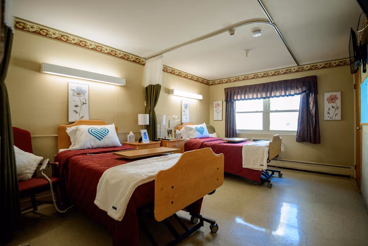 A nursing facility room with two hospital-style beds, each with a red quilted blanket and white pillow featuring a blue heart logo. Between the beds is a wooden nightstand with lamps, framed photos, and a tissue box. The room has beige walls with floral border trim near the ceiling, floral artwork on the walls, a large window with dark curtains, and a red armchair with a white pillow. The floor is a light-colored linoleum.