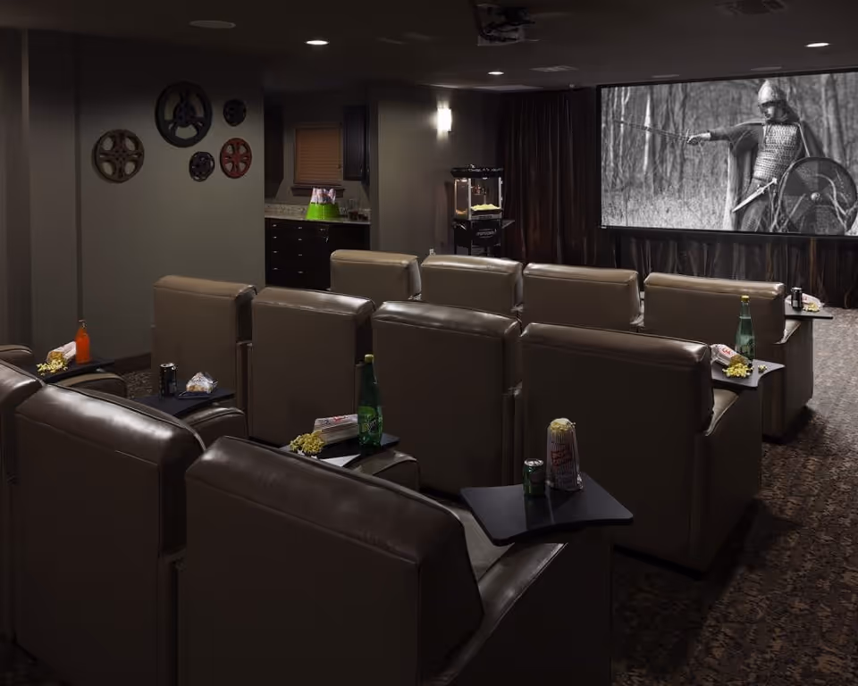 A cozy movie theater room with multiple rows of leather recliner chairs, each equipped with a small side table holding snacks and drinks. A large screen at the front displays a black and white scene of a medieval warrior in armor holding a sword and shield. The room has dim lighting, decorative film reels on the wall, and a popcorn machine near the back.