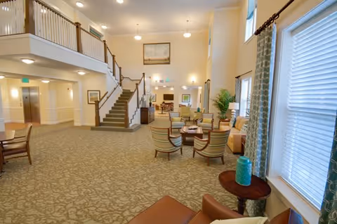 Spacious and well-lit senior living facility common area with a high ceiling, carpeted floor, multiple seating arrangements including armchairs and sofas, a staircase leading to an upper level, large windows with curtains, and decorative plants and artwork.