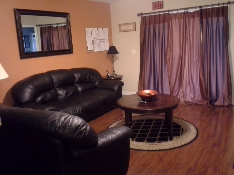 A cozy living room with a black leather sofa and armchair, a round wooden coffee table with a decorative bowl on top, a large mirror on the wall, a side table with a lamp, and purple curtains covering a window or door. The floor is wooden with a circular rug under the coffee table.