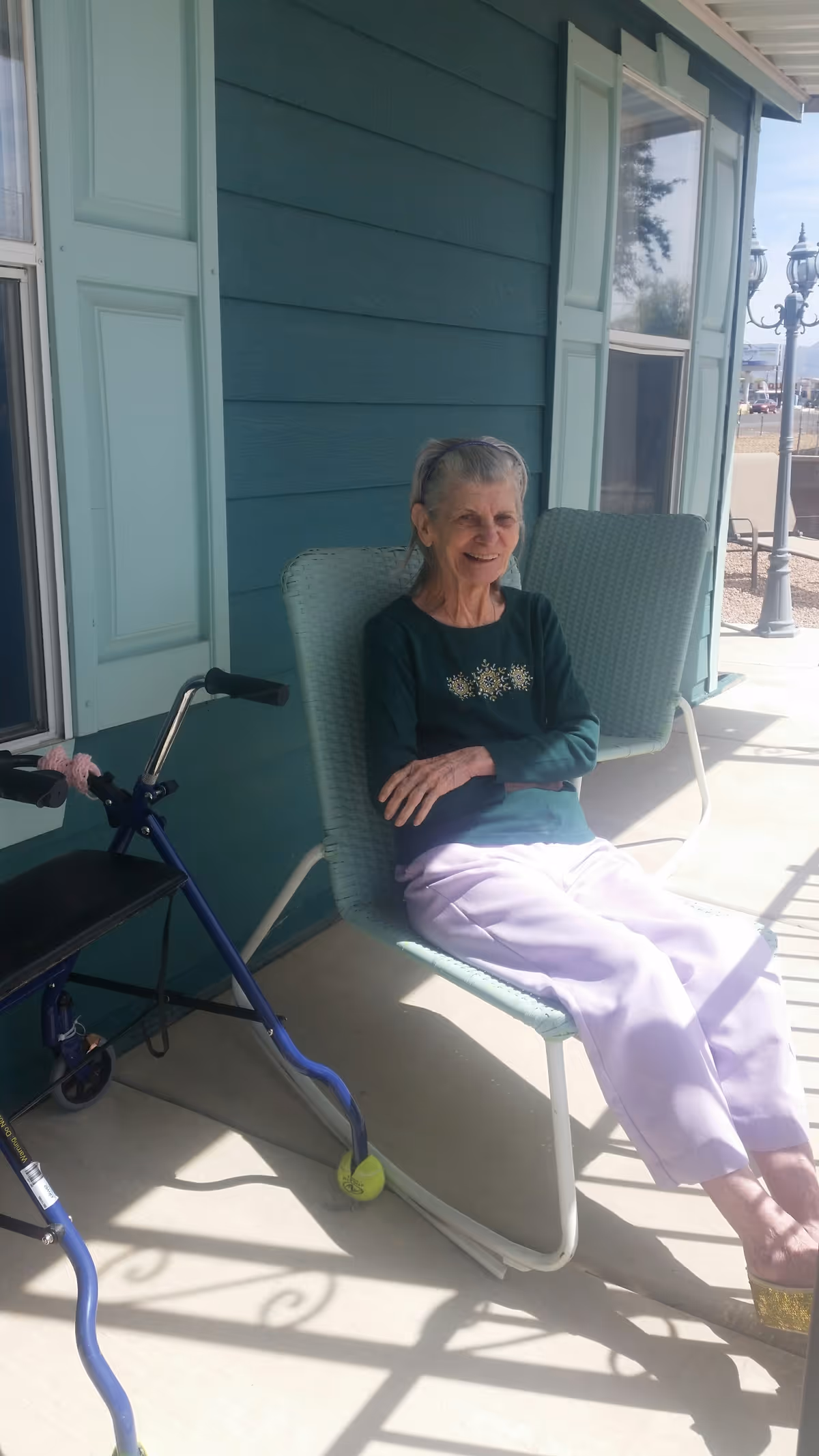 An elderly woman with gray hair is sitting on a light green outdoor chair on a porch. She is wearing a dark green long-sleeve shirt with a floral design and light purple pants. Next to her is a blue walker with tennis balls on the back legs. The porch has teal siding with windows and shutters, and there is a street lamp visible in the background.