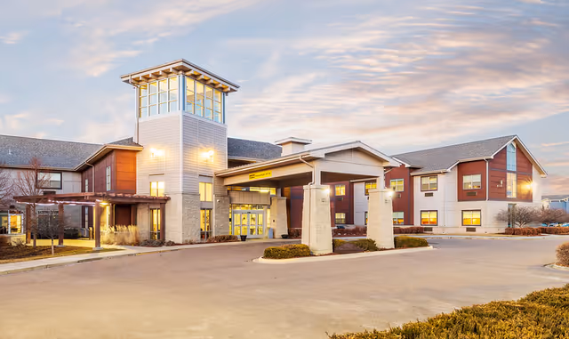 Exterior view of Coal Creek Post Acute and Assisted Living facility during sunset, showing a modern two-story building with a covered entrance supported by stone pillars and large windows.