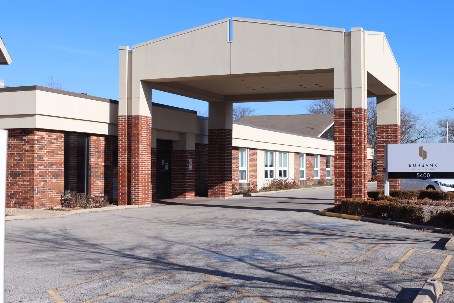 Front entrance canopy and driveway of Burbank Rehabilitation Center with brick columns and a sign showing the facility name and address.