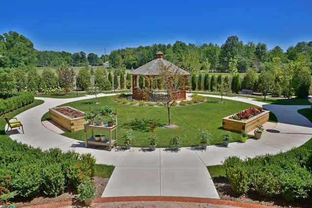 A well-maintained outdoor garden area with a circular concrete pathway surrounding a wooden gazebo in the center. The garden features raised flower beds with plants and flowers, small trees, bushes, and benches along the pathways. The sky is clear and blue, and there are trees and greenery in the background.