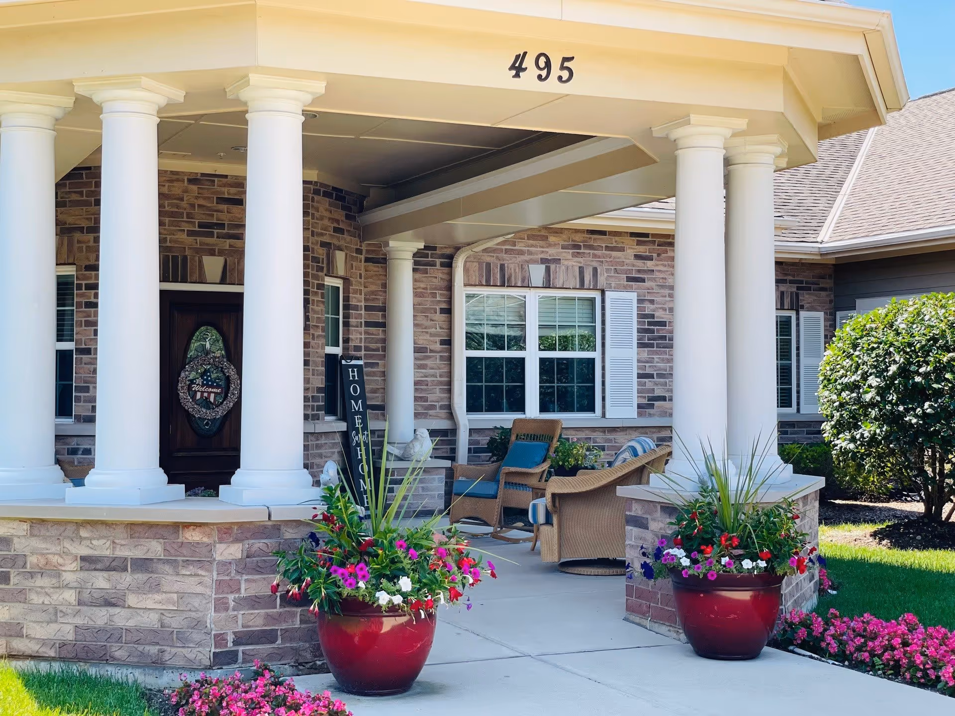 Front porch of a brick building with white columns and the number 495 displayed above. The porch has wicker chairs with cushions, a welcome sign, and large red flower pots with colorful flowers on either side of the walkway. Green bushes and a well-maintained lawn surround the area.