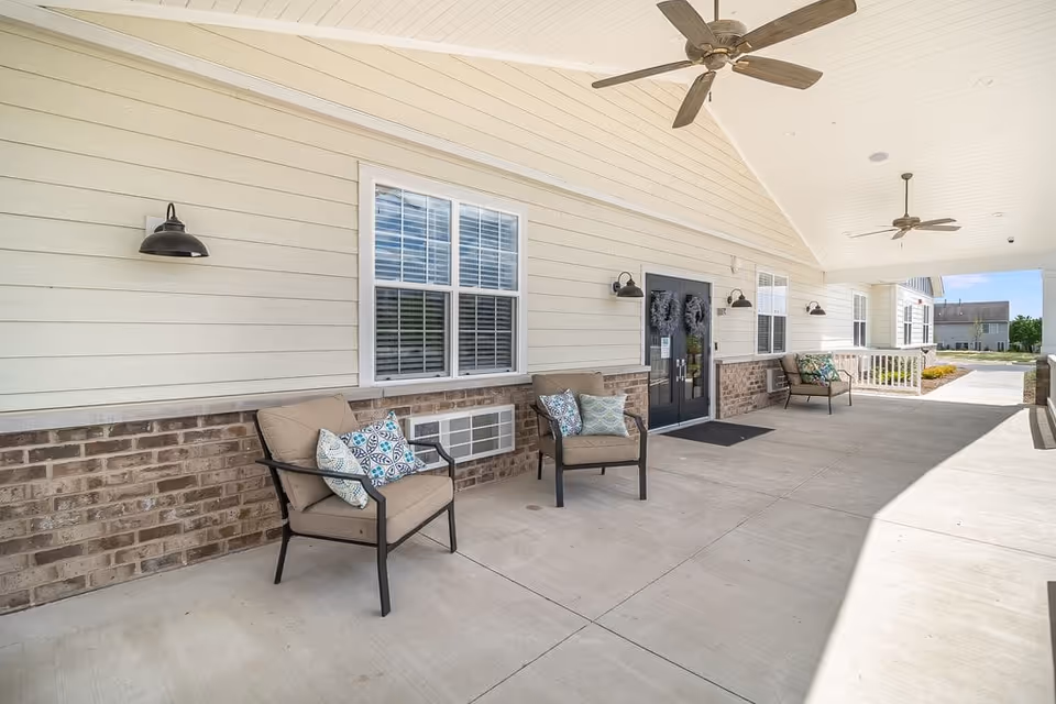 Covered outdoor patio area with beige cushioned chairs adorned with decorative pillows, mounted wall lights, ceiling fans, and double glass doors leading inside a building with light-colored siding and brick accents.