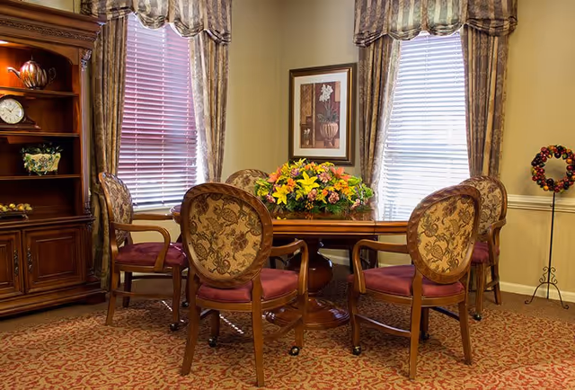 Warmly decorated communal dining area with a round wooden table, upholstered chairs, and a floral centerpiece in front of draped windows.