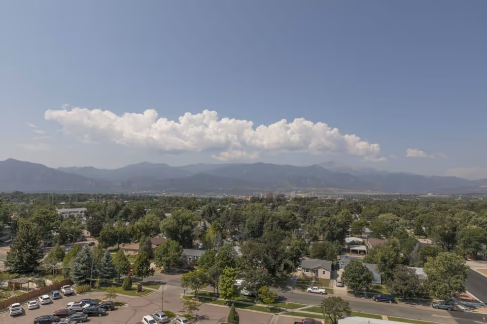 A wide aerial view of a tree-lined suburban neighborhood with parked cars and distant mountains under a partly cloudy sky.
